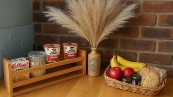 Una mesa de madera con un fondo de pared de ladrillo, que presenta una cesta de frutas, un florero con plumas de plumero y dos latas de Quaker Oats.