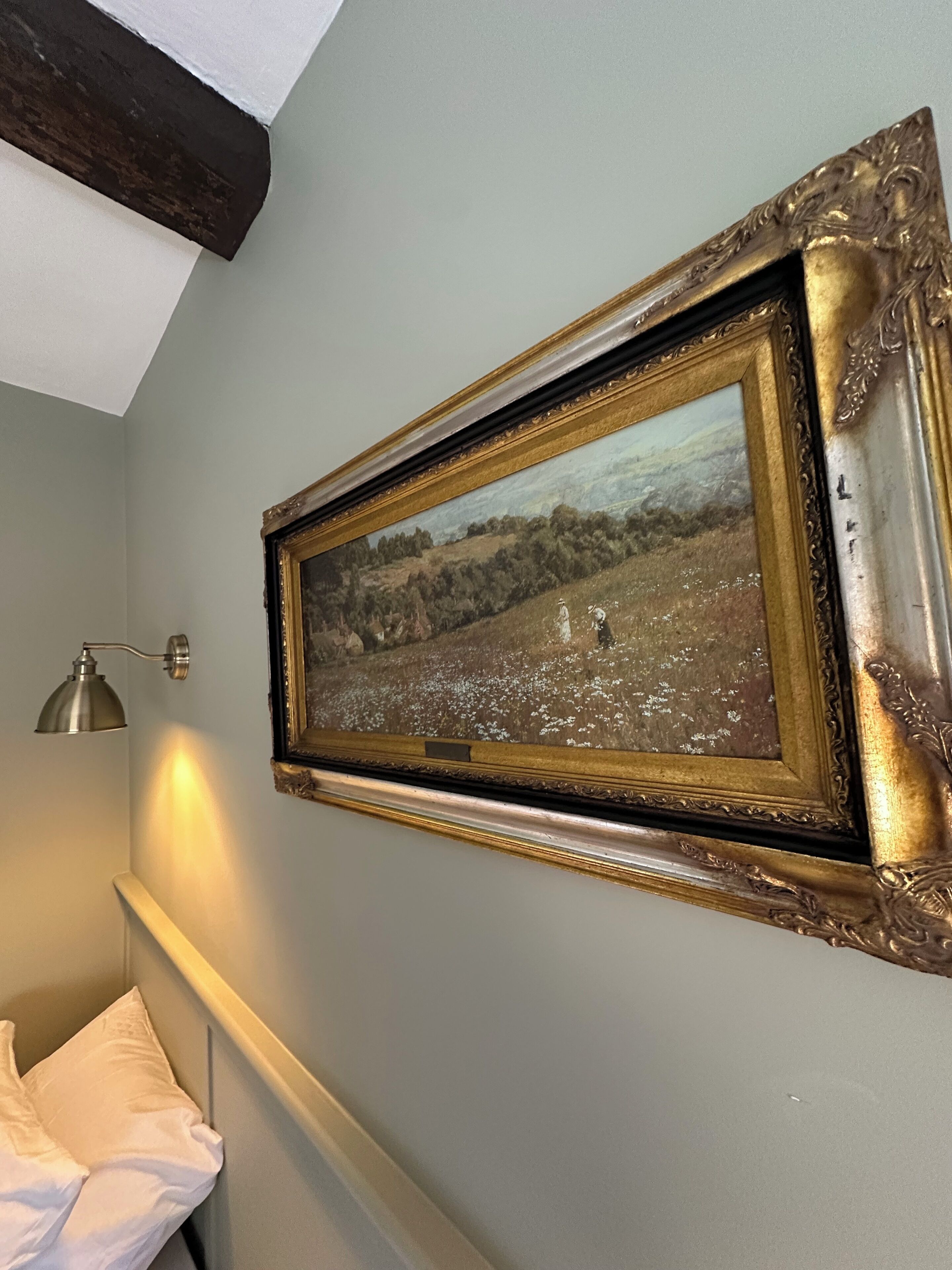 Un tableau représentant un paysage, encadré et accroché au mur, une lampe de chevet et un oreiller blanc sur un lit.