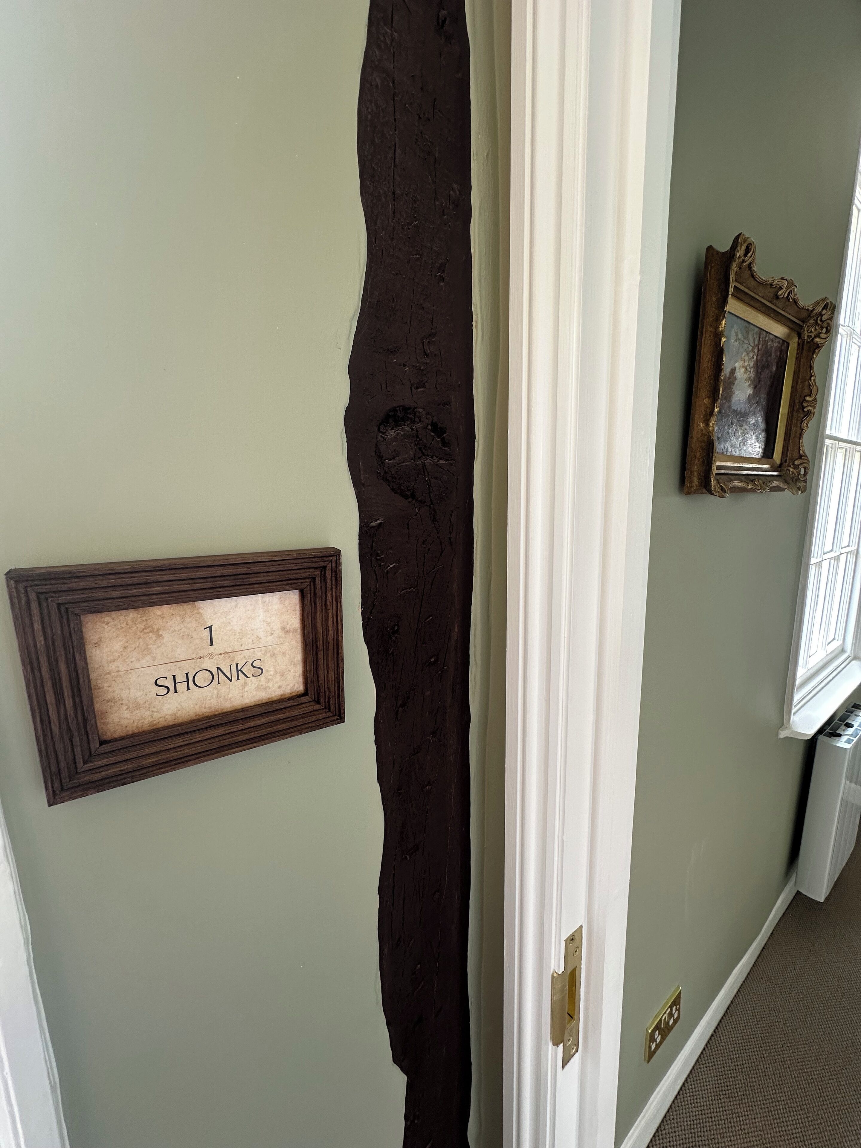 Un couloir avec un mur à la texture sombre, un cadre de porte, une enseigne encadrée portant l’inscription « 1 SHONKS » et un cadre photo.
