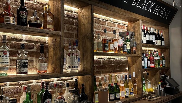 Zona de bar con un letrero que dice 'Welcome to The Black Horse', estantes con varias botellas de licores y una barra con vasos y una caja registradora.