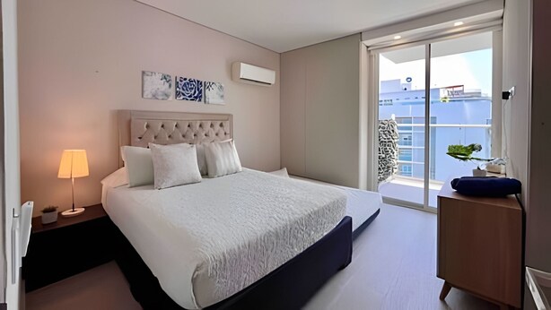A modern hotel room with a large bed, a nightstand, a lamp, a dresser, and a balcony with a view of the city. - Edificio Martinique  (Cartagena)