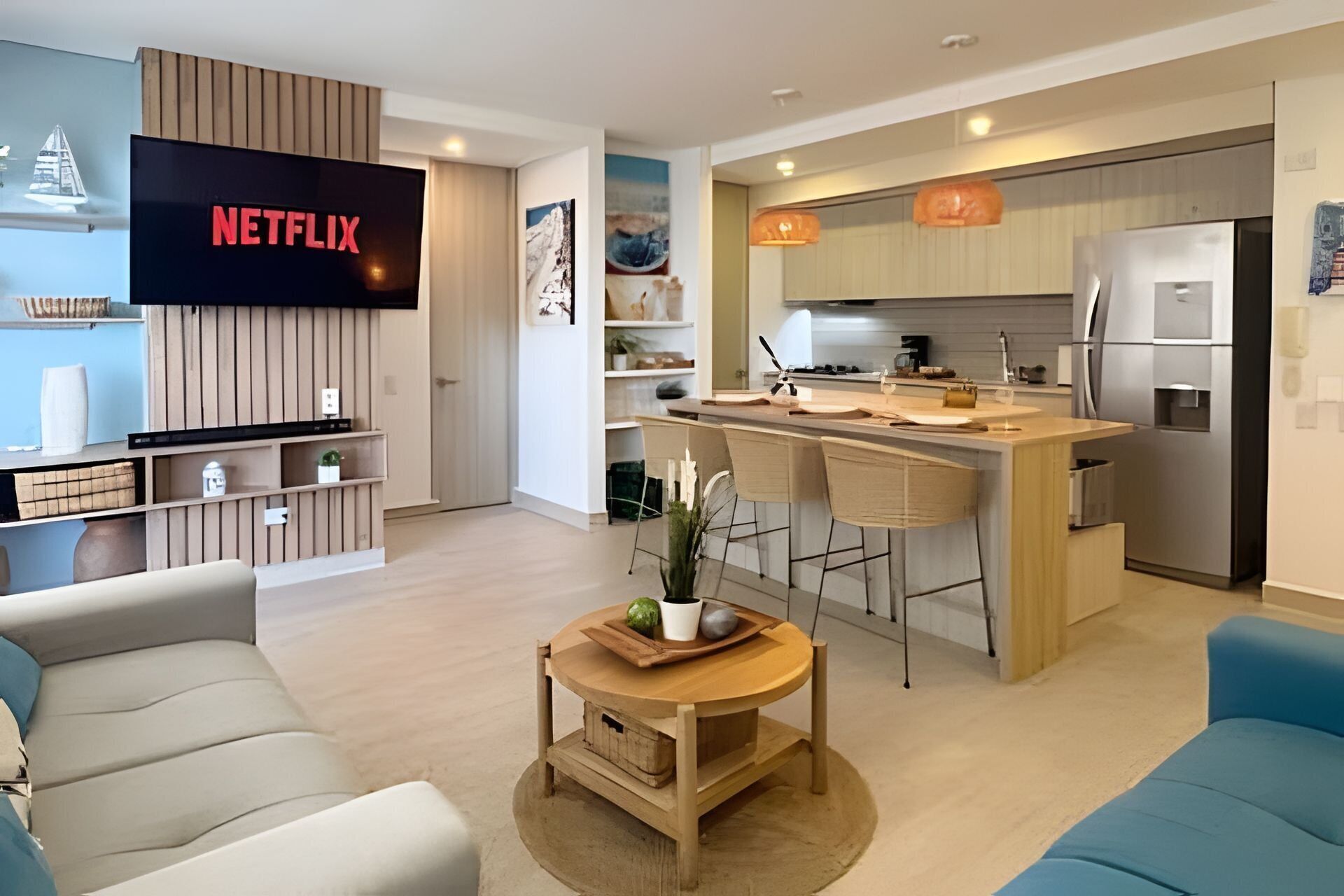 Una sala moderna con un televisor de pantalla plana mostrando 'NETFLIX', una cocina con refrigerador de acero inoxidable y una zona de comedor con mesa de madera.