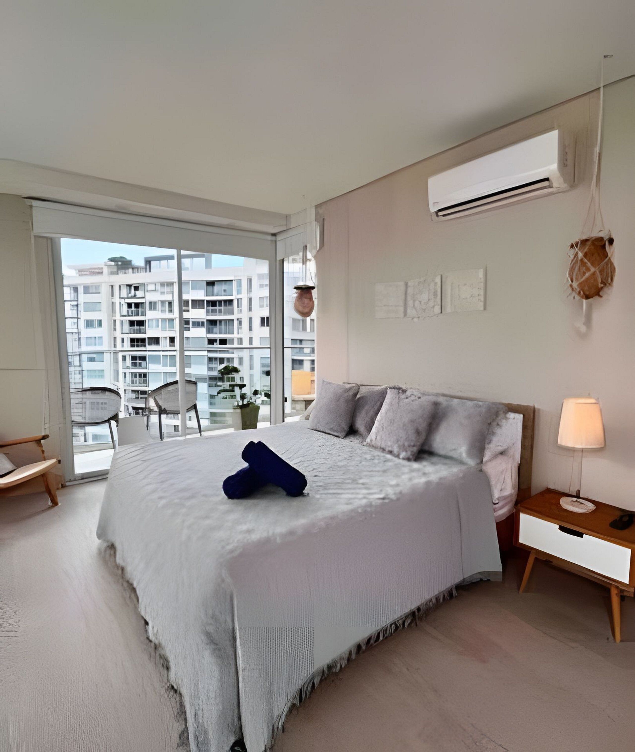 A bedroom with a large bed, a nightstand, a lamp, a hanging plant, and a view of a cityscape through a sliding door.