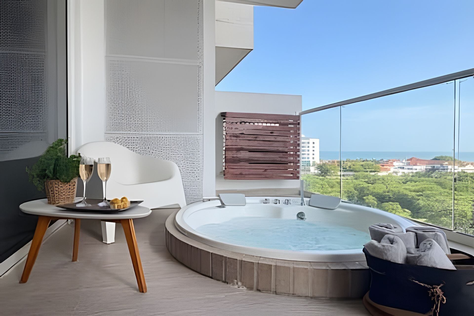 Un balcón moderno con un jacuzzi, una mesa con vasos y pasteles, y vistas a la ciudad.