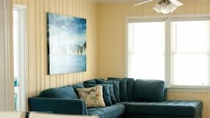 A living room with a sectional sofa, a ceiling fan, a window with blinds, and a painting on the wall.