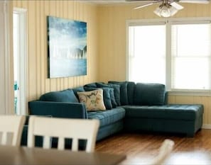 A living room with a sectional sofa, a ceiling fan, a window with blinds, and a painting on the wall.