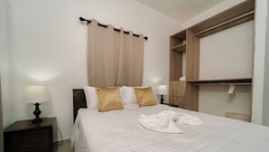 Een net opgemaakt bed met een witte quilt en twee gouden kussens, een bedlampje en een houten nachtkastje.