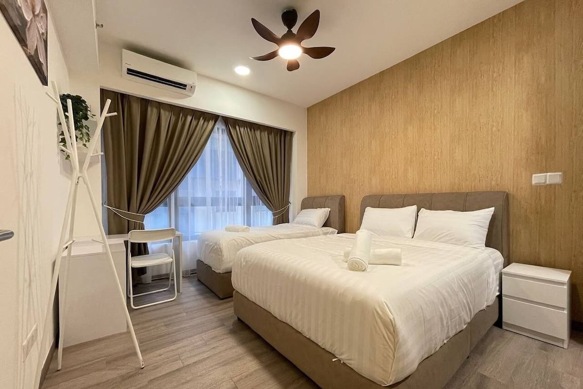 A hotel room with two beds, a desk, a chair, a standing fan, and an air conditioning unit.