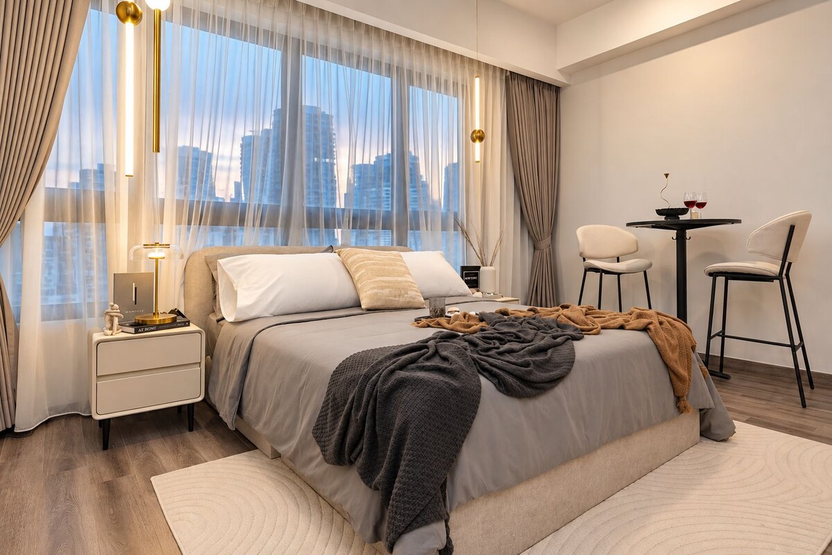 Un dormitorio moderno con vista a la ciudad, una cama con múltiples almohadas, mesitas de noche y un pequeño rincón de descanso con sillas y una mesa.