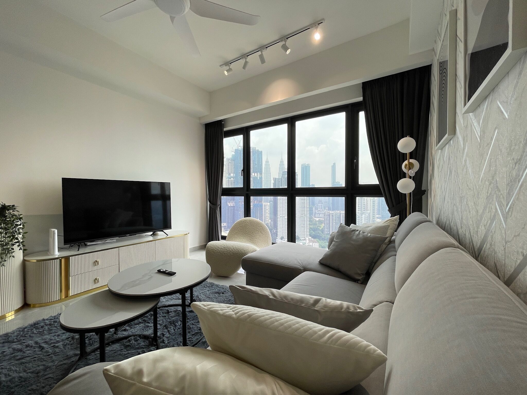 A modern living room with a large flat-screen TV, a round coffee table, a sofa, and a view of a city skyline through large windows.