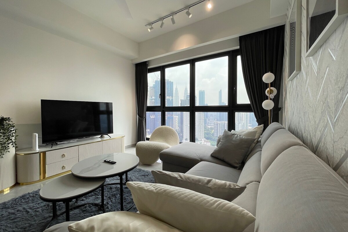 Una sala de estar moderna con un televisor de pantalla plana grande, una mesa redonda de centro, un sofá y vistas a un horizonte urbano a través de ventanas amplias.