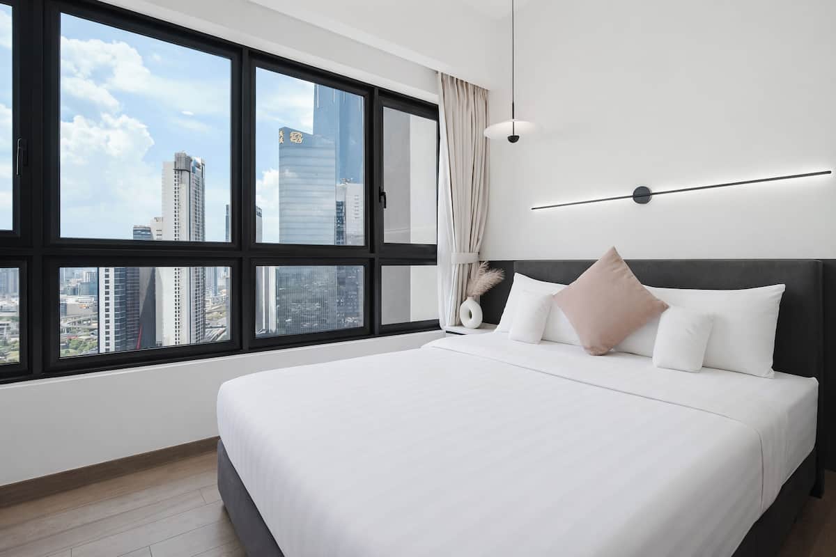 A hotel room with a large window, a bed with white bedding, a dark headboard, and a modern light fixture.
