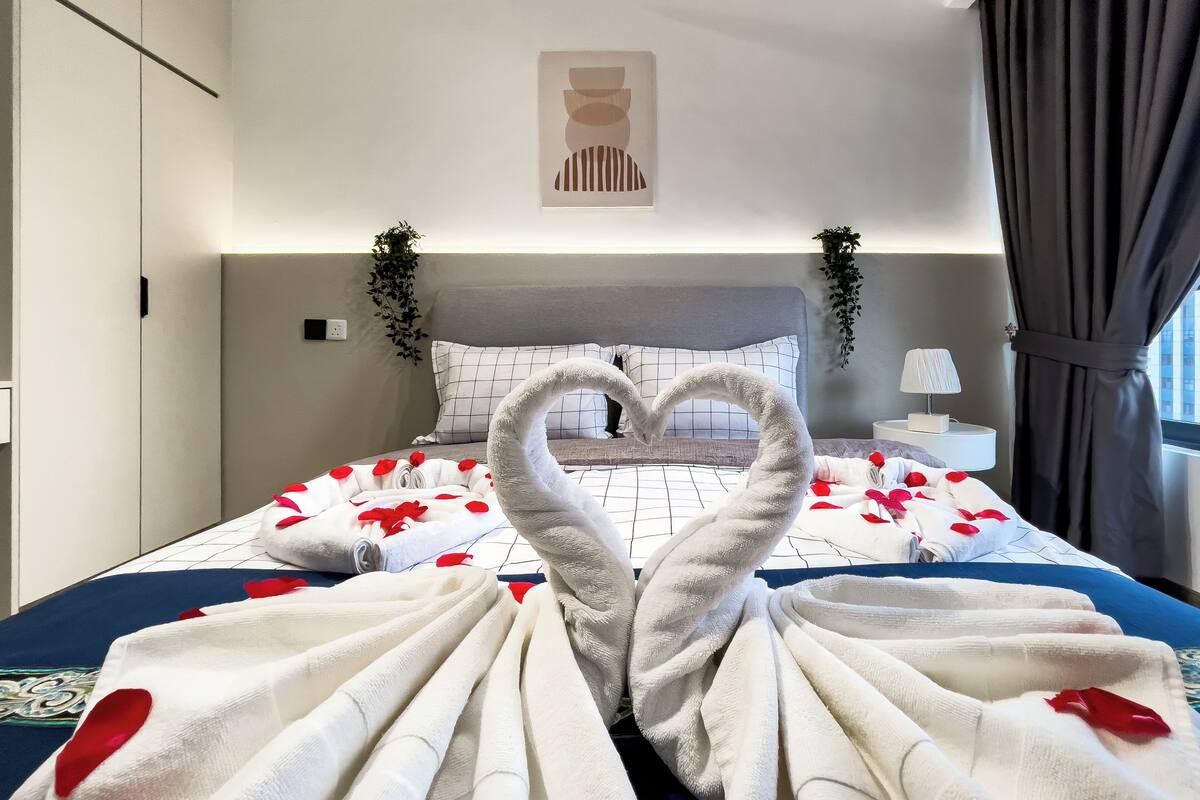 A hotel room with a bed featuring a heart-shaped towel arrangement, a bedside plant, and a view of the city through the window.