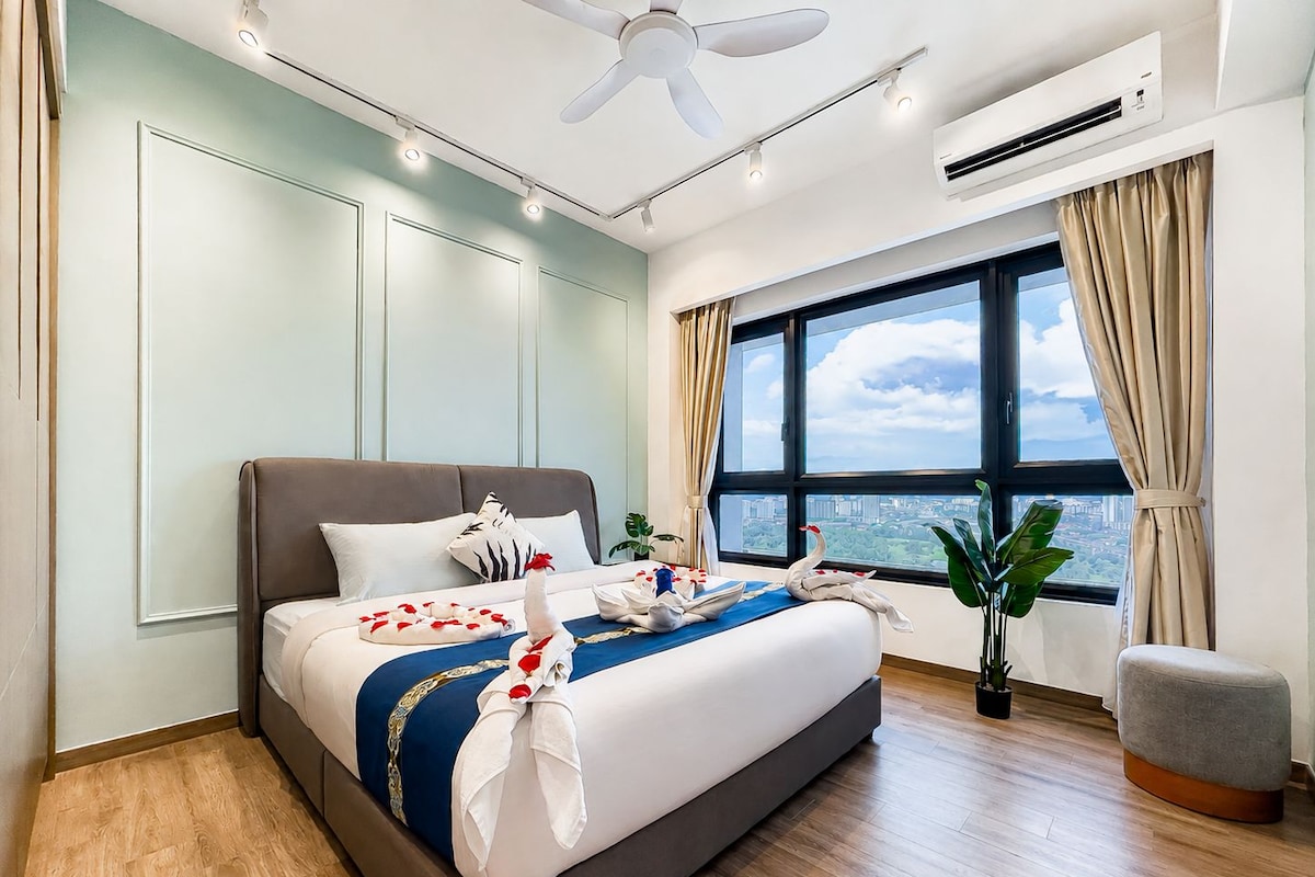 A hotel room with a large bed, a ceiling fan, an air conditioning unit, a window with curtains, and a view of the city.