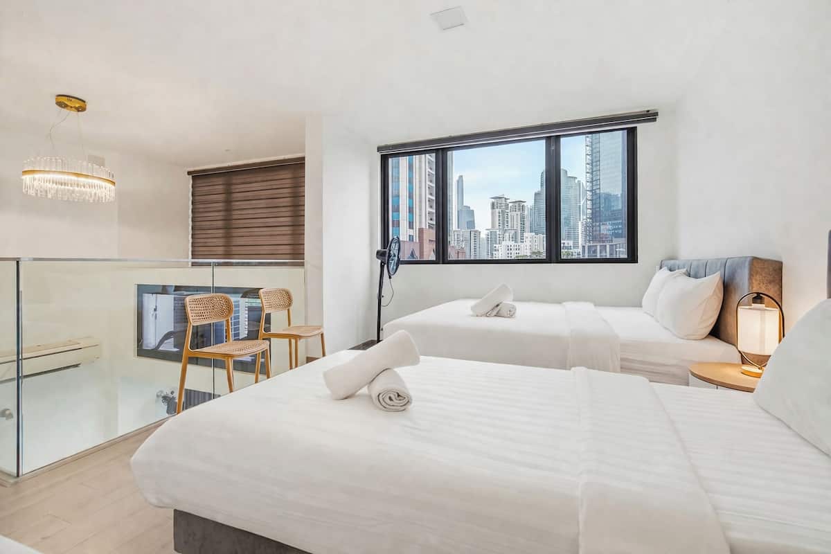 A modern hotel room with two beds, a city view, a glass partition, and a chandelier.