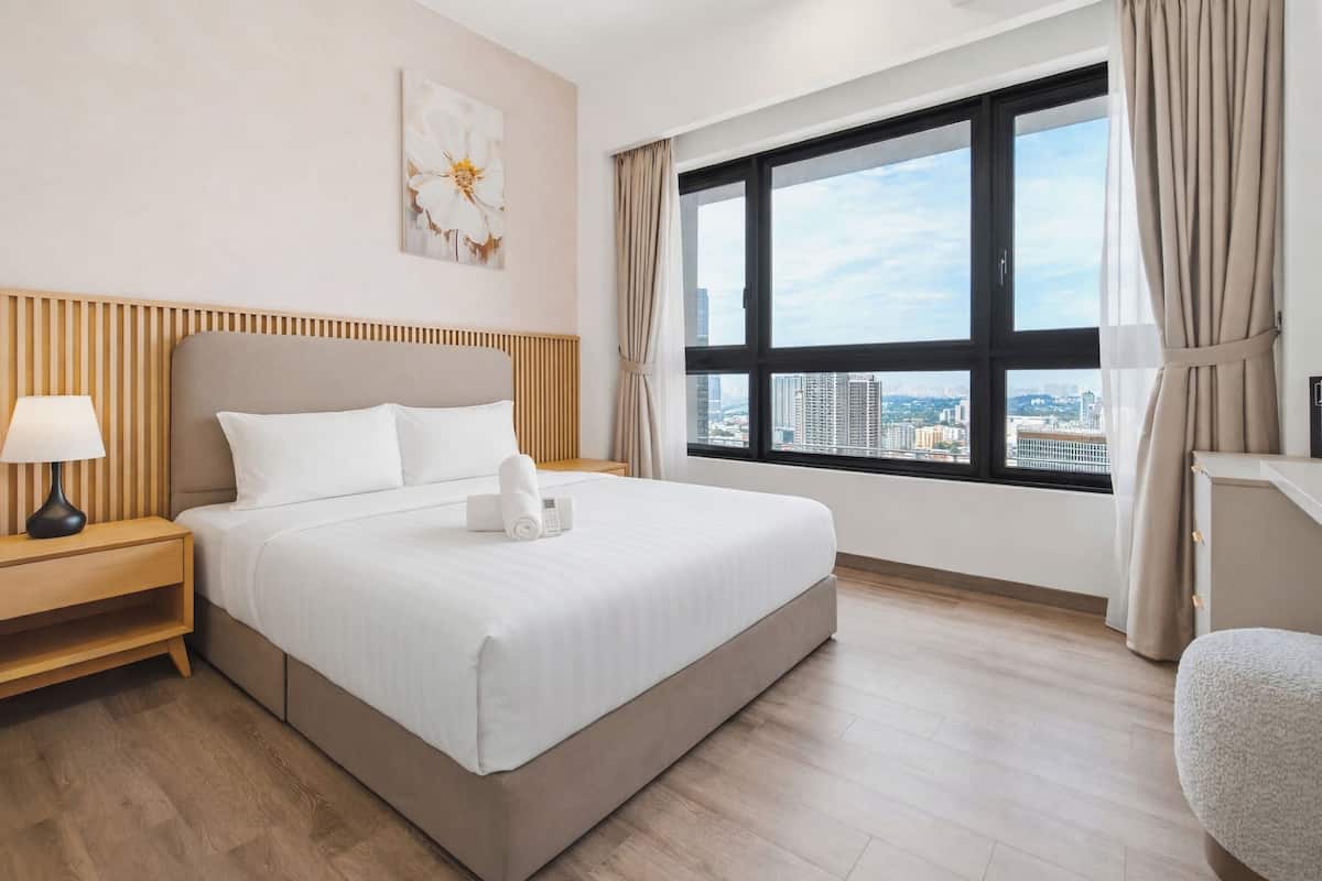A hotel room with a large bed, a bedside table with a lamp, a city view through a large window, and a painting on the wall.