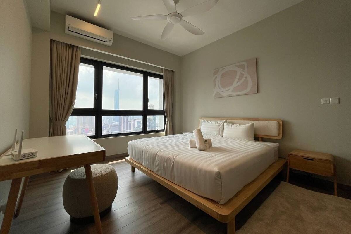Een hotelkamer met een bed, bureau, stoel, airconditioning, plafondventilator, raam met gordijnen en uitzicht op de stad.