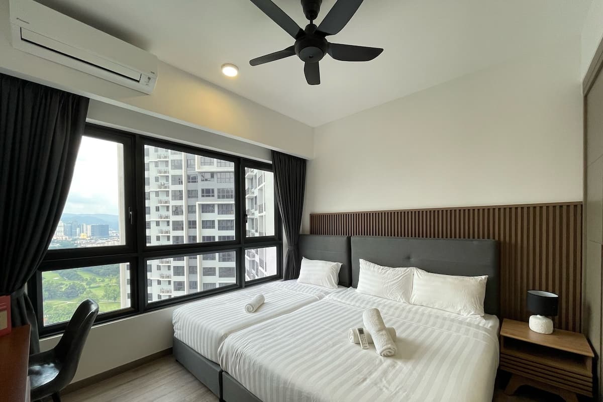 A hotel room with a large bed, a ceiling fan, a window with a city view, and a bedside table with a lamp.