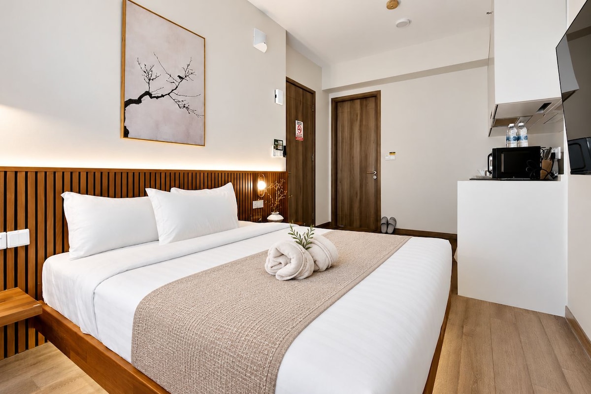 A hotel room with a bed, a wooden headboard, a wall-mounted artwork, a door, and a kitchenette area.