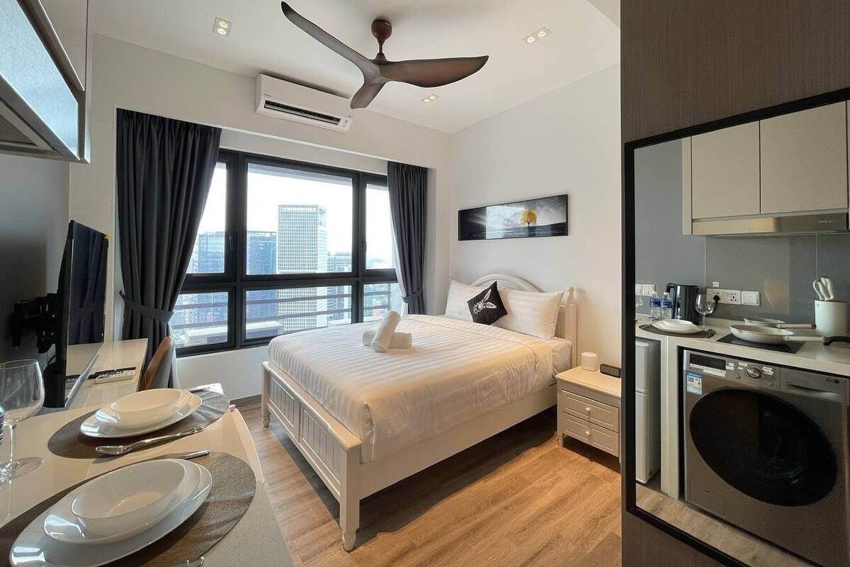 A modern hotel room with a large bed, a dining area, a kitchen, and a view of the city.