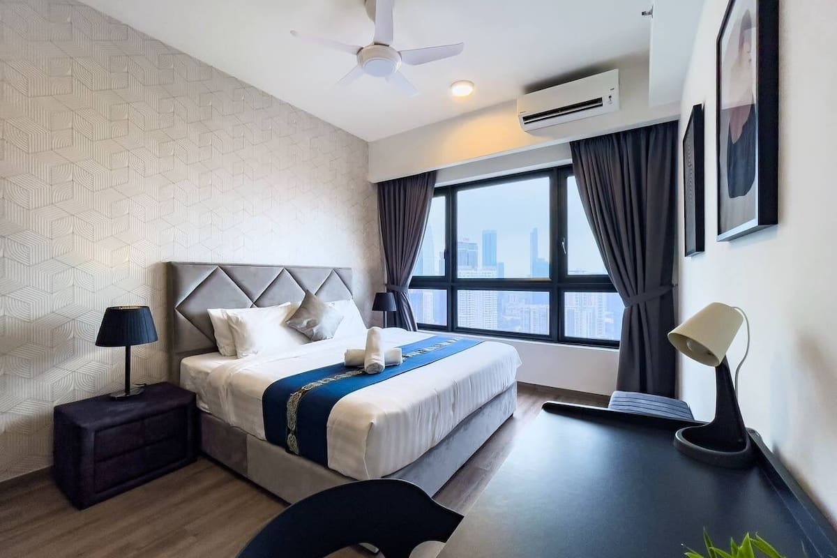 A modern hotel room with a large bed, a desk with a lamp, a chair, and a view of the city through the window.