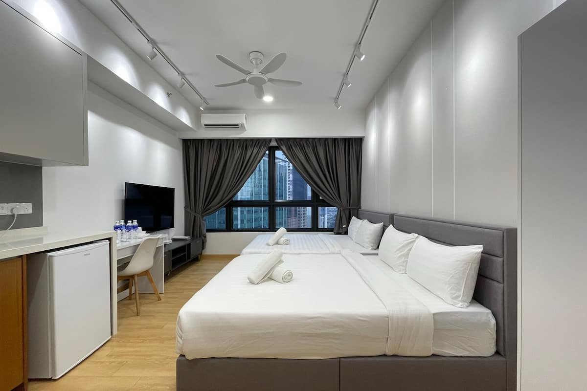 A modern hotel room with a large bed, a desk with a computer, a flat-screen TV, and a view of the cityscape through large windows.