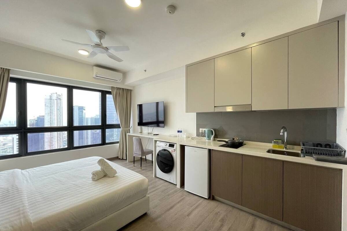 A modern hotel room with a bed, a washing machine, a kitchenette, and a large window with a city view.