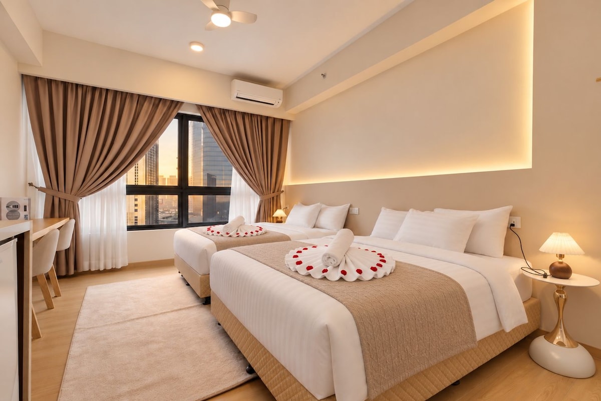 A hotel room with two beds, a desk, a chair, a desk lamp, and a view of the cityscape.