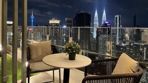 A balcony with a round table and two chairs, overlooking a cityscape with illuminated skyscrapers, including the Petronas Towers.