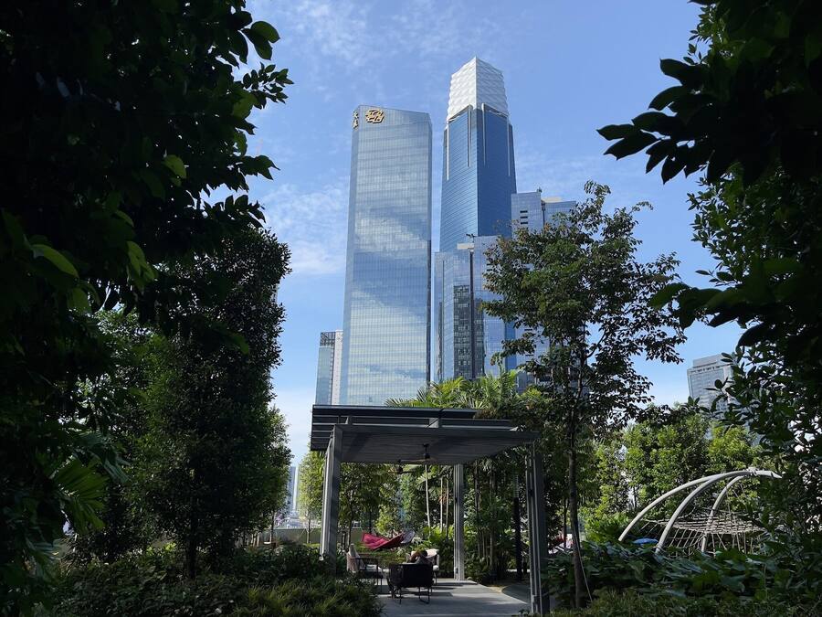 A modern urban park with a pavilion and seating area, surrounded by lush greenery and tall buildings in the background.
