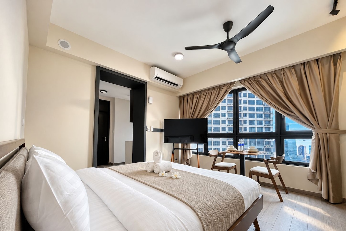 A hotel room with a bed, a TV, a ceiling fan, a window with curtains, and a view of buildings.