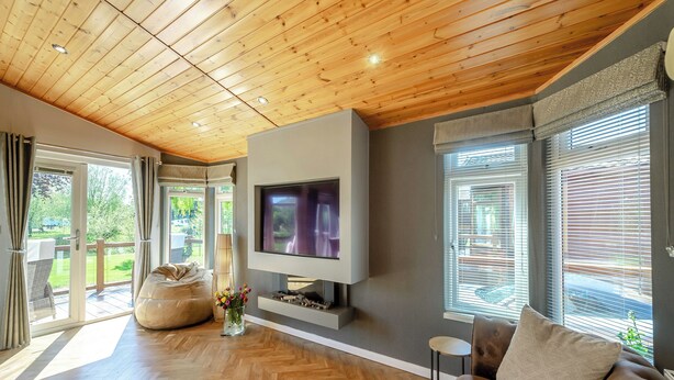 A spacious living room with a wooden ceiling, a large flat-screen TV mounted on a grey wall, a brown leather sofa, and a herringbone-patterned wooden floor.