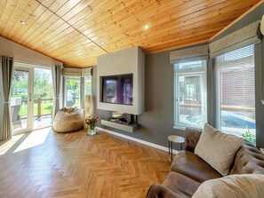 A spacious living room with a wooden ceiling, a large flat-screen TV mounted on a grey wall, a brown leather sofa, and a herringbone-patterned wooden floor.