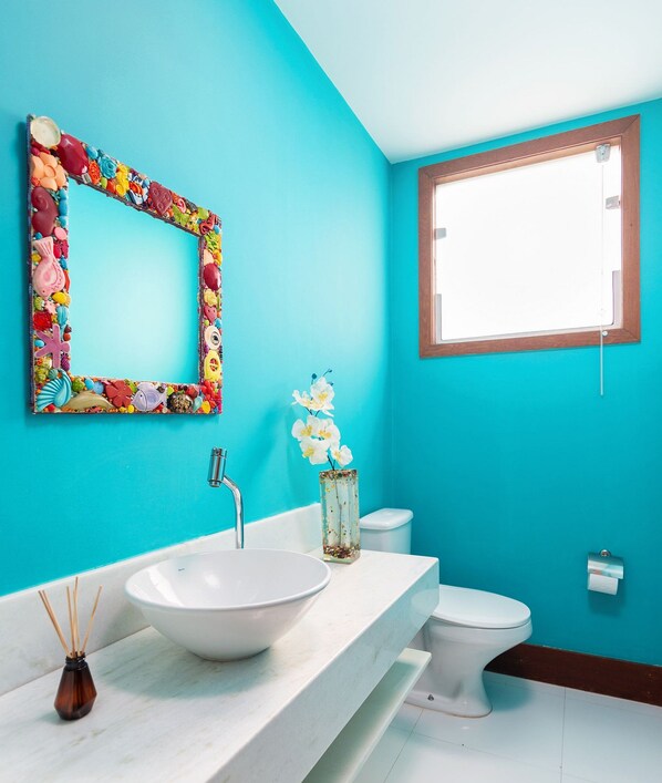 A bathroom with a white sink, a mirror with a colorful frame, a toilet, and a window with a wooden frame. - Bz33 Casa 4 suítes churrasqueira Geribá (Buzios)