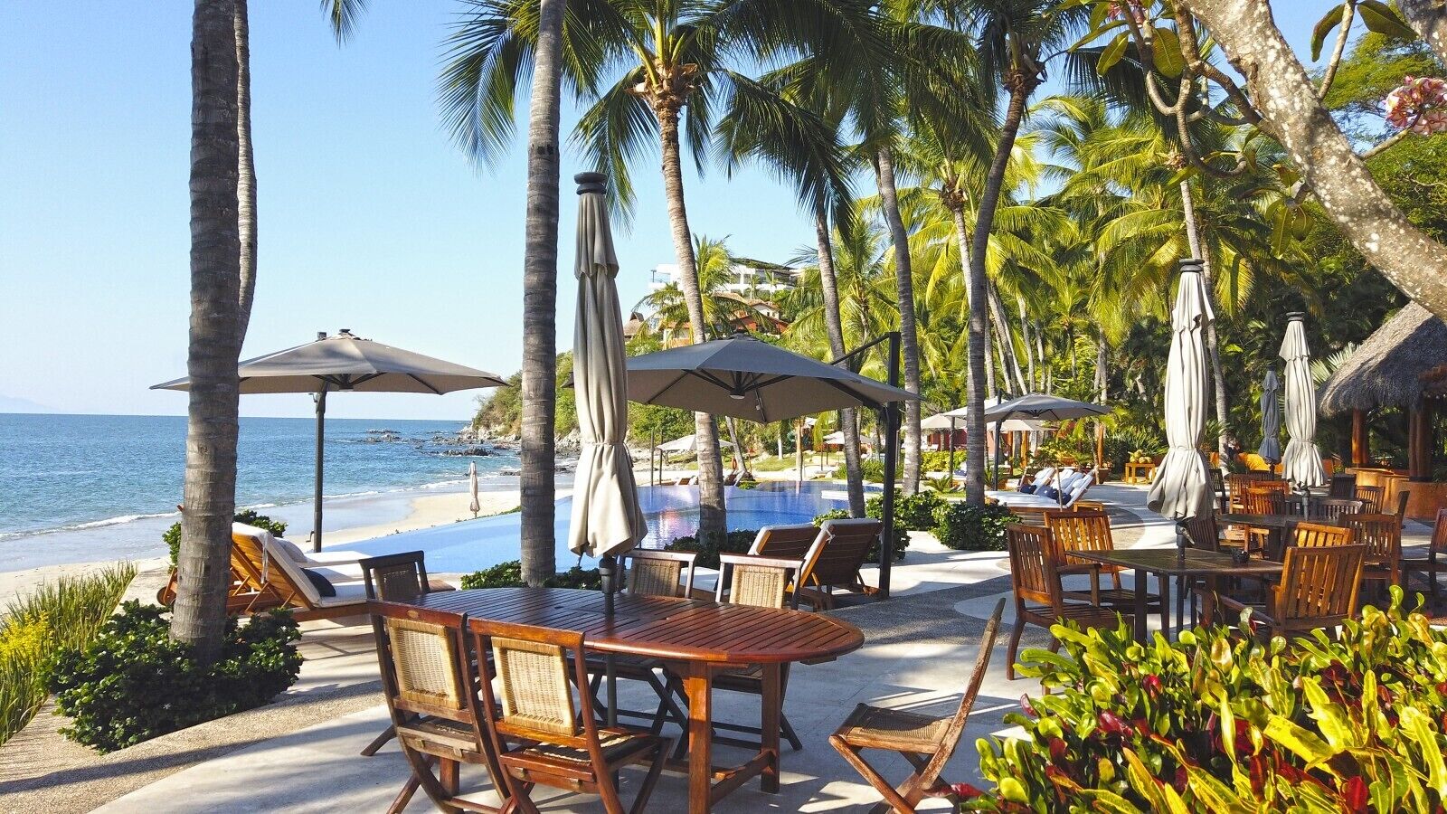 Un resort frente a la playa con terraza al aire libre, sombrillas y piscina.