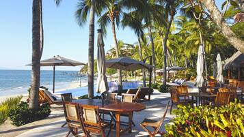 Un resort frente a la playa con terraza al aire libre, sombrillas y piscina.