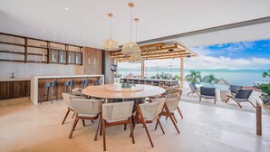 A spacious dining area with a round table and chairs, a kitchen with wooden cabinets, and a view of the ocean. - Staffed villa in gated community - Punta Mita Corridor (La Cruz de Huanacaxtle)