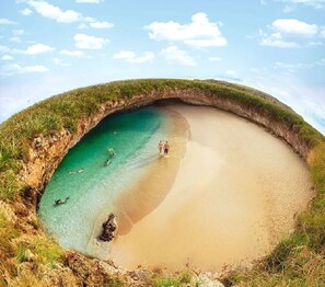 Une plage à l’eau turquoise et limpide, un rivage sablonneux et une petite entrée de grotte.