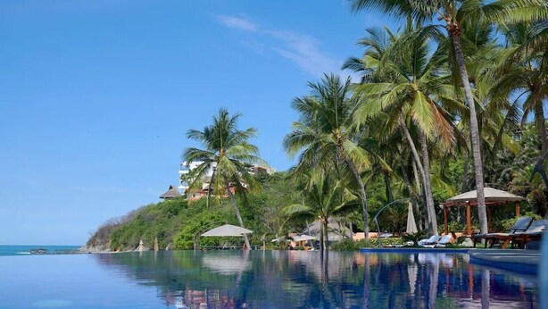 A pool with a clear reflection, surrounded by palm trees and a building with a thatched roof. - Staffed villa in gated community - Punta Mita Corridor (La Cruz de Huanacaxtle)