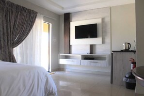 Habitación de hotel moderna con una cama grande, un televisor de pantalla plana montado en la pared, una kitchenette pequeña y una puerta corrediza de vidrio al exterior.