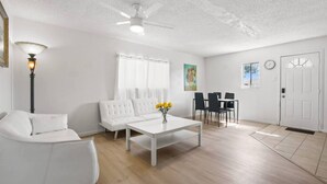 A living room with a white sofa, a coffee table with flowers, a dining area with chairs, a ceiling fan, and a white door.