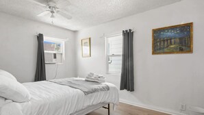 A bedroom with a bed, a ceiling fan, a window with curtains, a painting on the wall, and a white bucket.