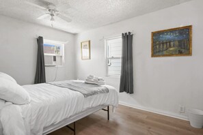Un dormitorio con cama, ventilador de techo, ventana con cortinas, un cuadro en la pared y un cubo blanco.