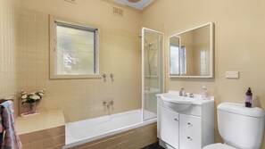 Ein Badezimmer mit einer Badewanne, einem Waschbecken und einer Toilette. Über der Badewanne befindet sich ein Fenster und über dem Waschbecken ein Spiegel.