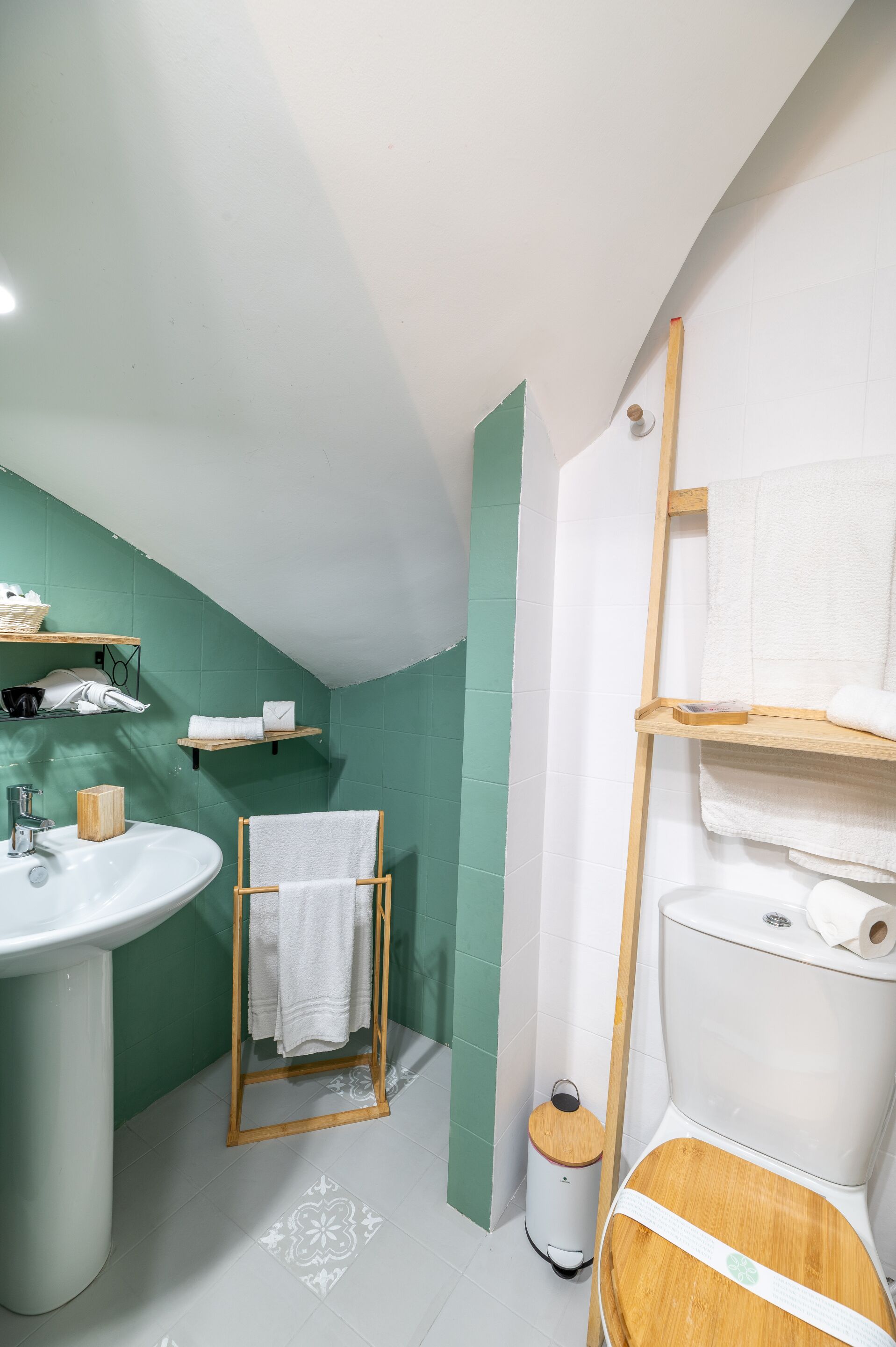 Une salle de bain avec un lavabo blanc, un meuble-lavabo en bois et des toilettes avec un abattant en bois. On y trouve des étagères avec des serviettes et une petite poubelle.
