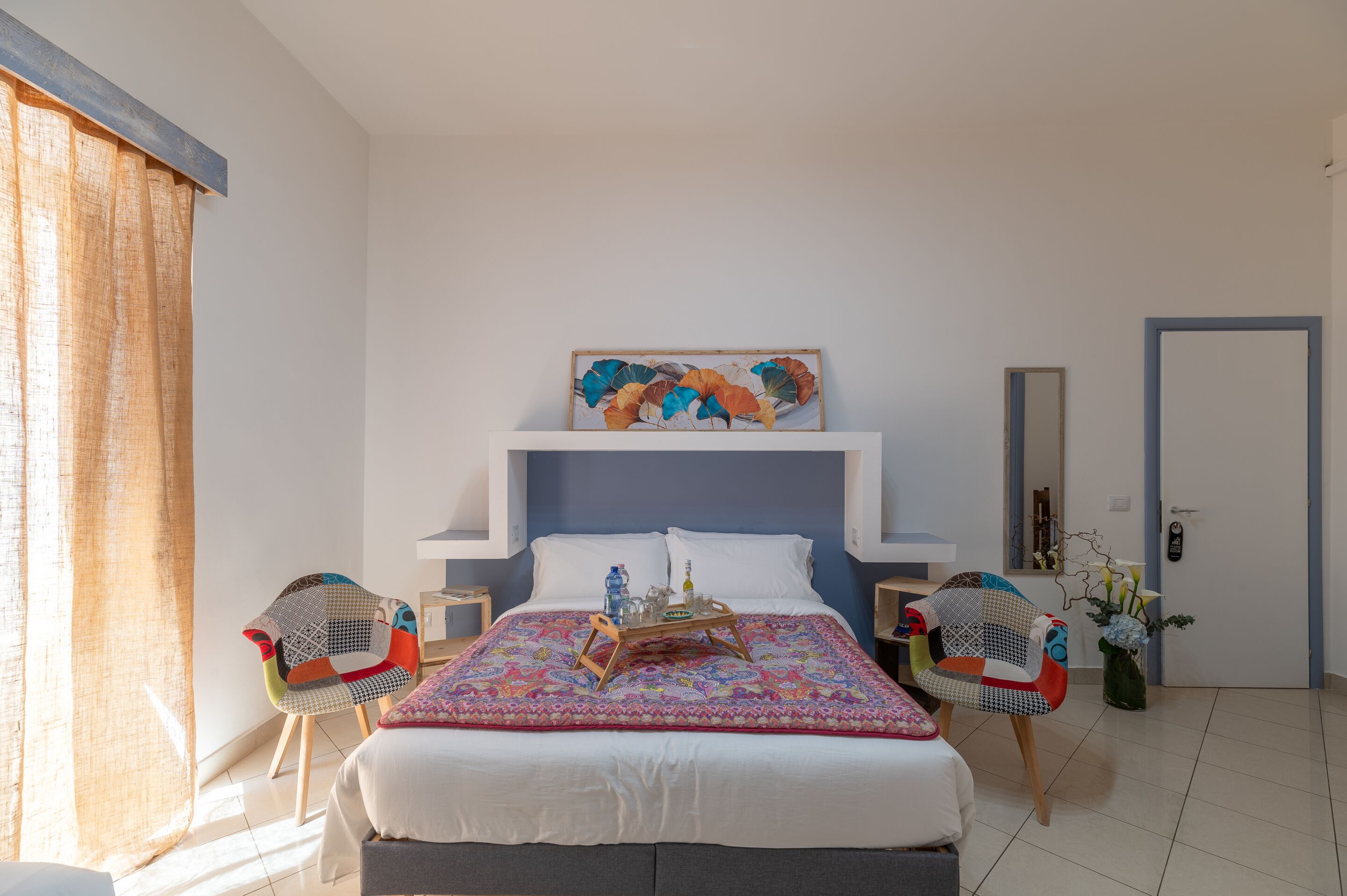 Een slaapkamer met een bed, twee stoelen, een nachtkastje, een spiegel en een schilderij.