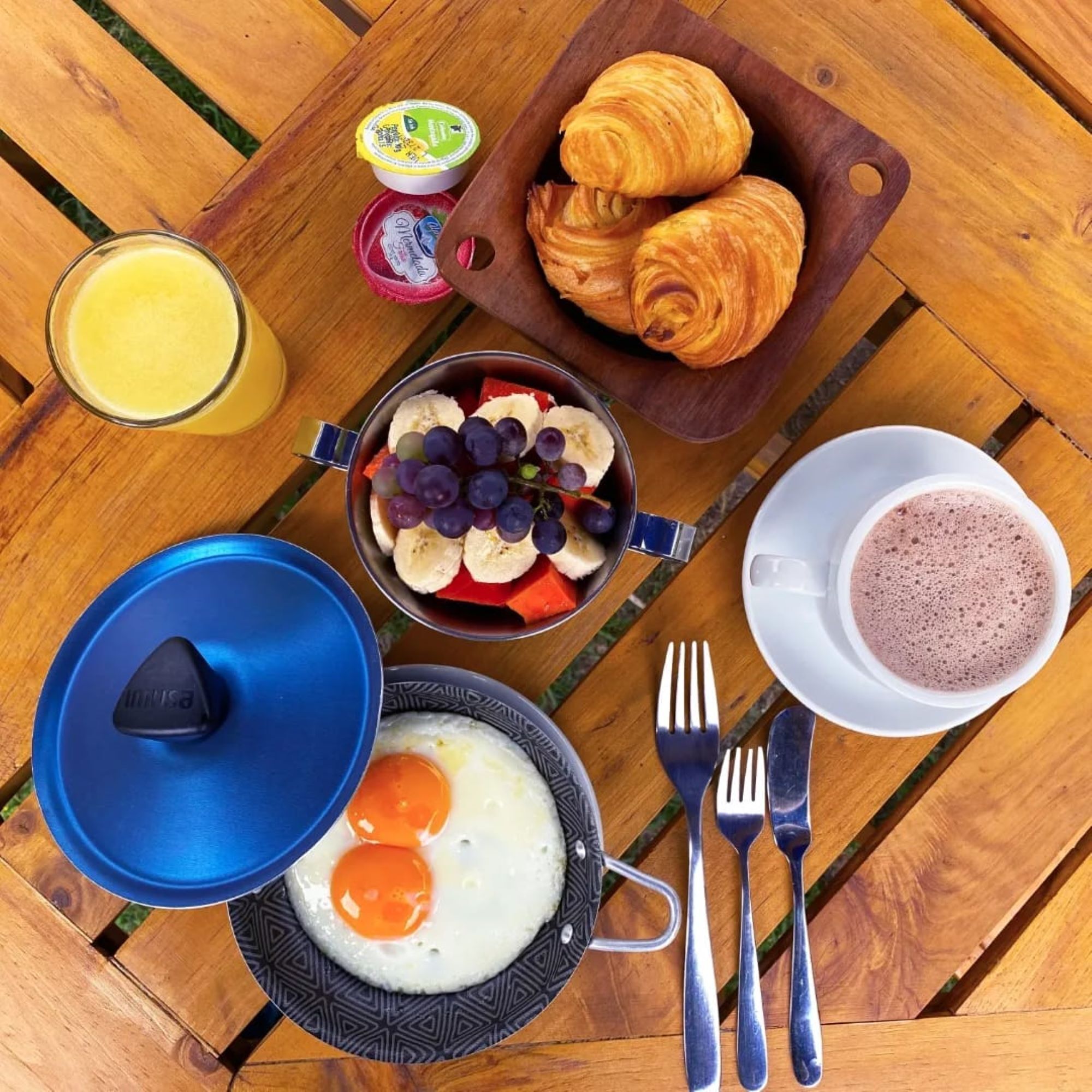 A breakfast setup with eggs, croissants, a bowl of fruit, a glass of orange juice, and a cup of coffee on a wooden table.