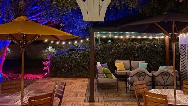 An outdoor patio with wooden flooring, a covered seating area with cushions, and string lights.