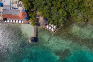 Vista aérea de un resort con un pasarela de madera que lleva a una estructura de techo de paja, un área de piscina con sombrillas y un mar turquesa cristalino.