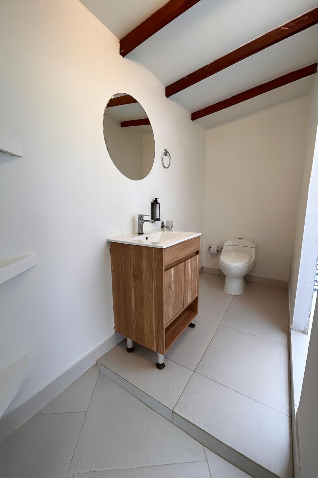 A bathroom with a wooden vanity, a round mirror, a toilet, and a white sink.